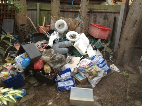 A pile of mixed household rubbish and waste materials placed on the ground in a backyard setting, with a wooden fence and trees in the background. The debris includes plastic bottles, cardboard boxes, paper, a cardboard box, a gray plastic flexible duct, a black plastic container, and various unidentifiable rubbish items. Some items appear to be discarded household items such as a white plastic bucket, a red plastic storage bin, and a brown cardboard box. The waste is haphazardly arranged, covering part of a dirt surface, with irregular shapes and a variety of textures, including smooth plastic, rough cardboard, and crumpled paper. The environment suggests an outdoor area in an urban or suburban location, where a rubbish removal service from Commercial Waste Removal Catford could assist in clearing such waste.