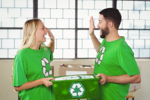 The image shows two waste management workers in a well-lit industrial or warehouse setting, both wearing bright green T-shirts with a white recycling symbol printed on the front. The female worker, with blonde hair, is smiling and touching her face as she faces the male worker, who has a dark beard and short hair. He is smiling back and giving a high-five gesture. The male worker is holding a green plastic recycling bin with a white recycling logo on the front, which is positioned in front of them. Behind them, there are large windows with a grid pattern allowing natural light to fill the space, and some cardboard boxes are visible in the background, indicating a waste collection or recycling environment. The scene reflects a positive interaction related to waste management, fitting for a commercial waste removal service operating in Catford, with the setting emphasizing environmental sustainability and recycling efforts in a professional context.