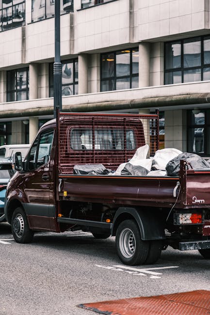 A small burgundy flatbed truck parked on a city street in front of a modern office building with large glass windows and light-colored stone cladding. The vehicle's cargo bed is filled with various ba