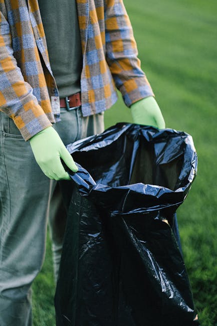 A waste collection worker wearing a high-visibility orange vest and dark trousers is seen emptying a large, red commercial waste collection truck parked beside a roadside. The truck has a metal rear l
